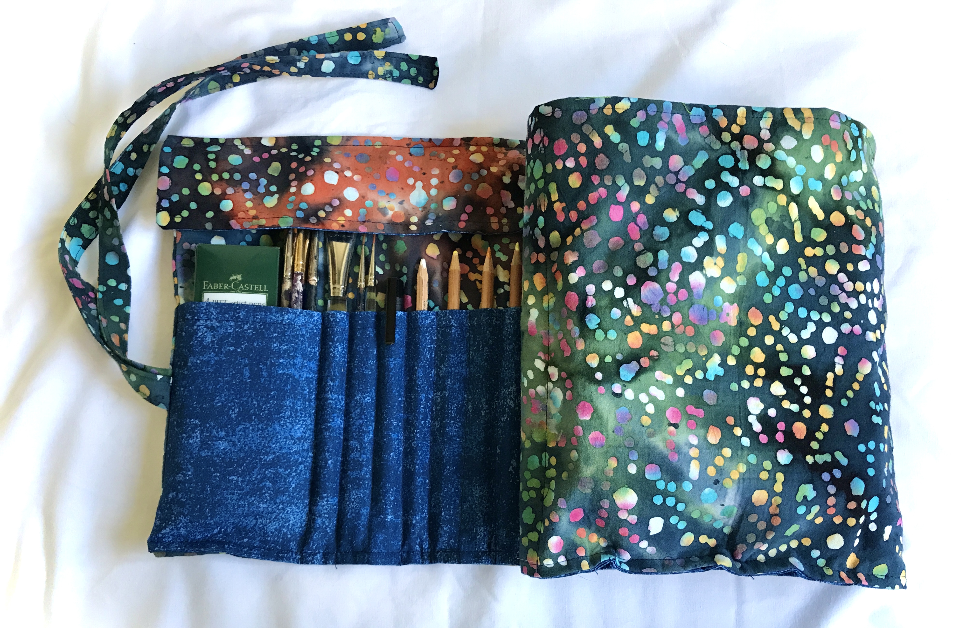 A fabric art supply roll with a blue, orange, and green batik pattern. The roll is partially open, revealing art supplies such as paint brushes, pencils and a pack of ink pens. A protective flap covers the top of the top of the brushes. The roll has a blue interior with pockets to hold the supplies.