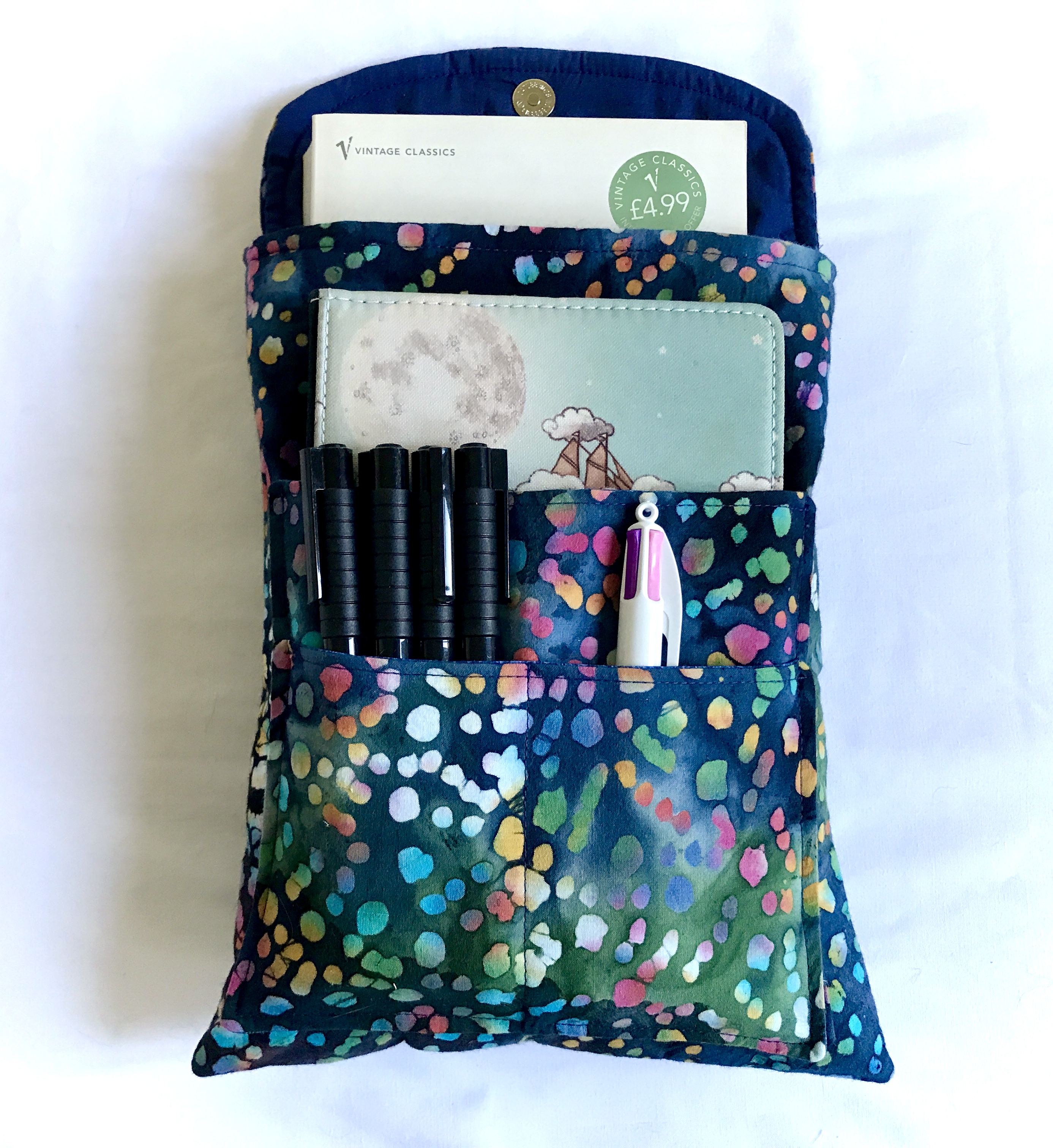 A dark blue book sleeve with colourful speckles of white, yellow, blue, green, orange, red, and pink. The book sleeve has its flap open, showing a magnetic clasp. It has a book peaking out of the top of the main bag, a kindle, in its cover sitting, in one of the front pockets, and a variety of pens in the front two pockets.
